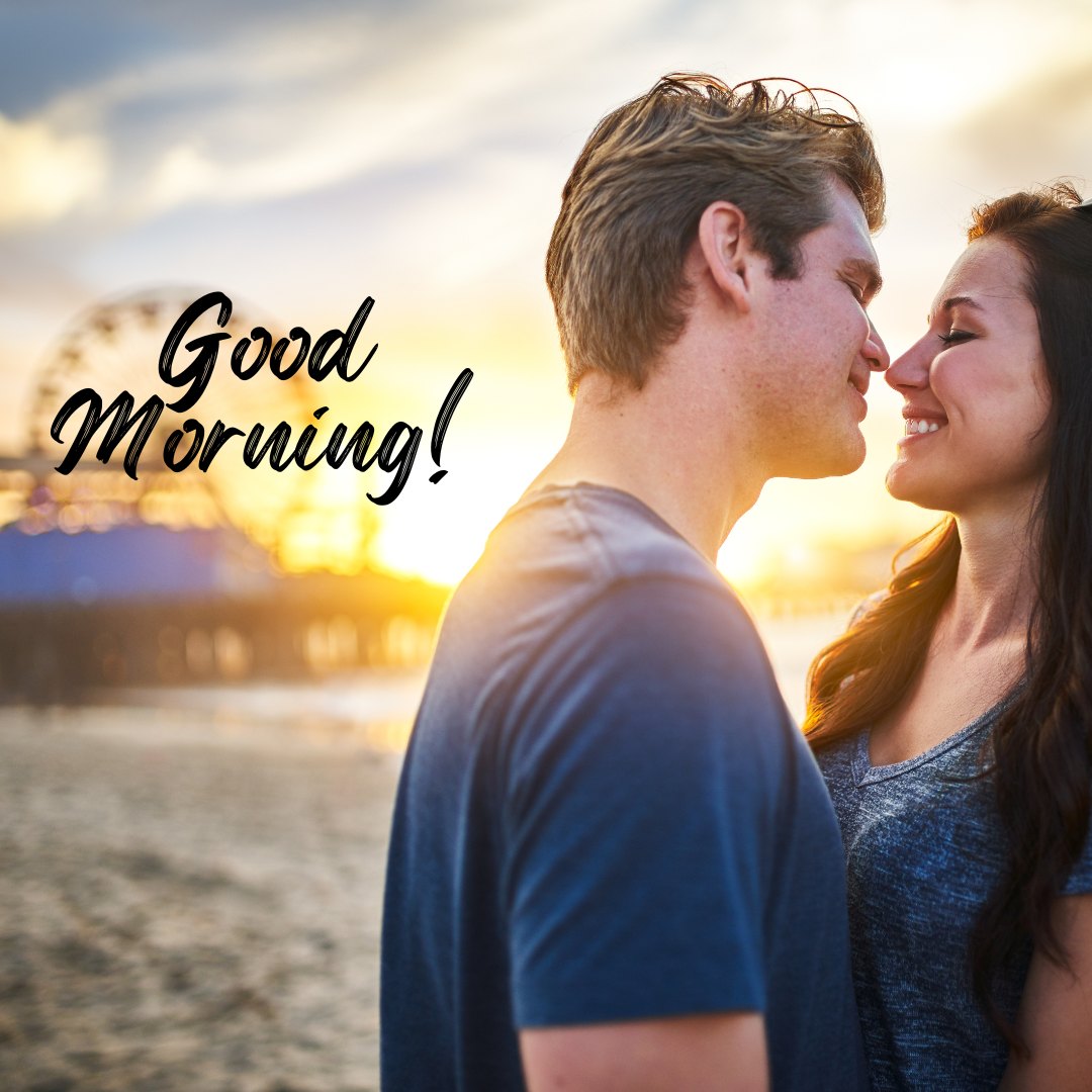 Good Morning Kiss Image featuring a couple exchanging a tender kiss by the seaside at sunrise, with a beautiful sky and a distant ferris wheel in the background.